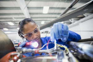 A female wearing safety glasses and gloves shining a torch at and holding an electrical wire that is part of a large piece of equipment in the foreground of the image.