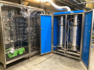 A metal cabinet with clear doors on the left showing an array of pipework, dials and other instrumentation. On the right, a solid metal cabinet painted blue with two large metal cylinders sitting vertically. A large pipe extends from the top of the blue cabinet