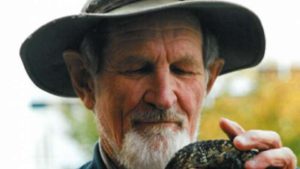 A elderly man in a hat holding and looking at a reptile
