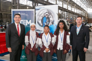 Three school girls standing between two men in suits in front of banners. they are standing in a very large shed with manufacturing and light industry activities taking place behind them.