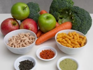 Red and green apples, brocolli and carrots with processed products derived from these fresh fruits and vegetables sitting in bowls in front of them.